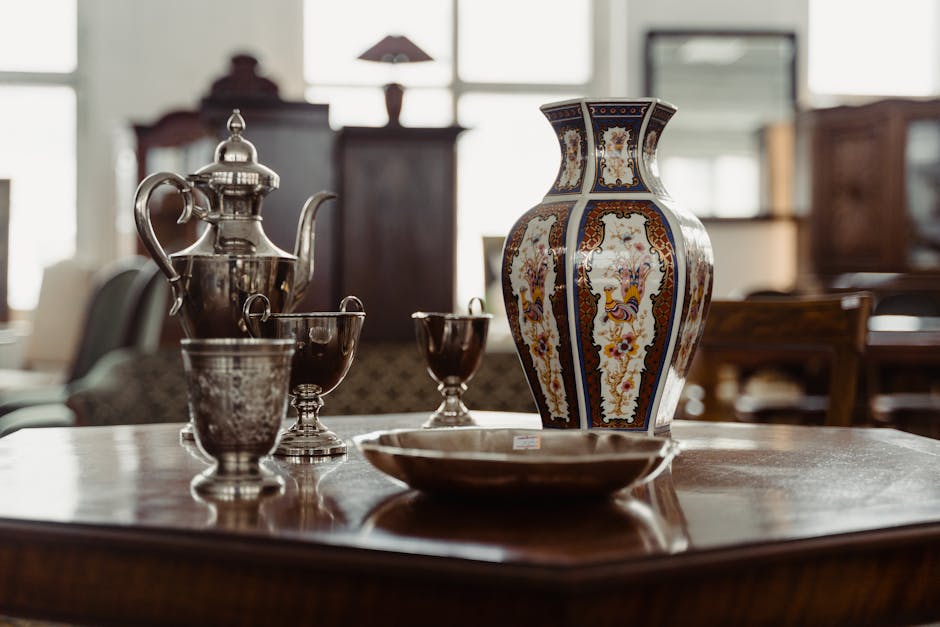 A woman wearing a light-colored face mask, a beige checked blazer, a patterned top, and jeans is inspecting or examining a set of white porcelain tea service placed on a wooden table inside an antique