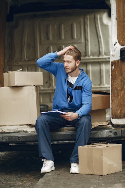 A young man with light brown hair and a beard, wearing a blue hoodie, dark blue trousers, and white sneakers, is sitting inside the back of a large moving van with the rear door open. He is holding a