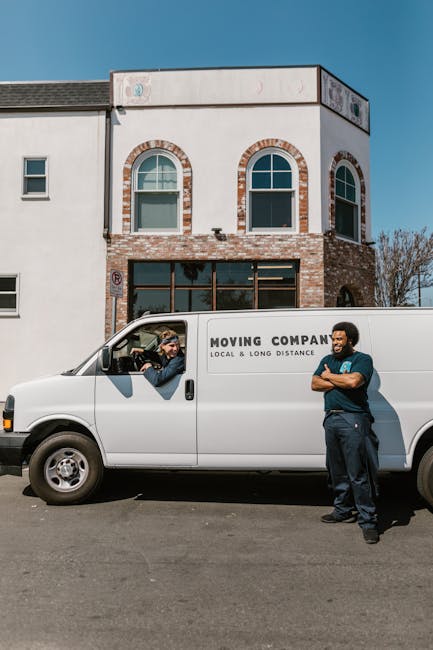 A young man with light brown hair and a beard, wearing a blue hoodie, dark blue trousers, and white sneakers, is sitting inside the back of a large moving van with the rear door open. He is holding a