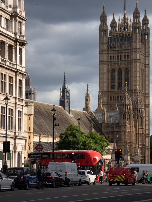 The image shows a busy street in Westminster with the iconic Palace of Westminster and Big Ben clock tower in the background under a bright blue sky with wispy clouds. Along the street, there are seve
